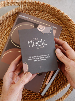 Hands holding a Fleck Deck prompt card over the Fleck Journal on a woven tray