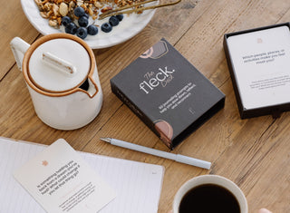 Open fleck deck, journal and pen on wooden table next to white teapot and bowl of blueberries and muesli